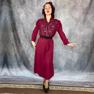 May include: A burgundy dress featuring a long-sleeved, embroidered bodice and a matching skirt. The dress is cinched at the waist with a black belt. The model is wearing black heels.
