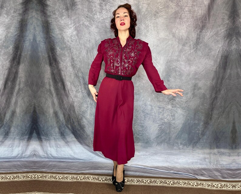 May include: A burgundy dress featuring a long-sleeved, embroidered bodice and a matching skirt. The dress is cinched at the waist with a black belt. The model is wearing black heels.