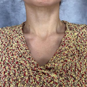 May include: Close-up of a textured knit sweater in shades of yellow, brown, and red. The sweater features a wide, folded collar and a V-neck design. The knit fabric has a speckled appearance.