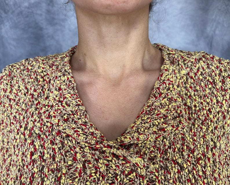 May include: Close-up of a textured knit sweater in shades of yellow, brown, and red. The sweater features a wide, folded collar and a V-neck design. The knit fabric has a speckled appearance.