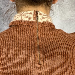 Pu&ograve; includere: Primo piano di un maglione lavorato a maglia color ruggine con colletto fantasia color crema. Il maglione presenta una cerniera a tutta lunghezza sulla schiena. La maglia ha una trama a coste.