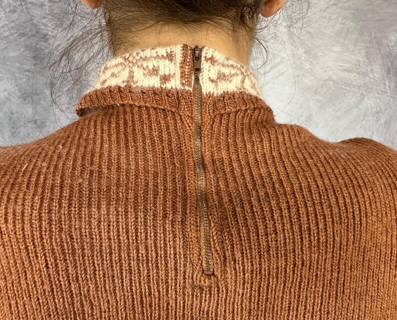 Pu&ograve; includere: Primo piano di un maglione lavorato a maglia color ruggine con colletto fantasia color crema. Il maglione presenta una cerniera a tutta lunghezza sulla schiena. La maglia ha una trama a coste.
