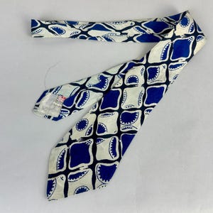 May include: A necktie featuring a repeating geometric pattern of dark blue rounded squares and white shapes with scalloped edges. The tie has a pointed end and a label on the underside near the narrow end, displaying a white background with blue.