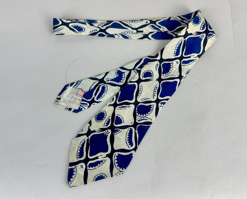 May include: A necktie featuring a repeating geometric pattern of dark blue rounded squares and white shapes with scalloped edges. The tie has a pointed end and a label on the underside near the narrow end, displaying a white background with blue.