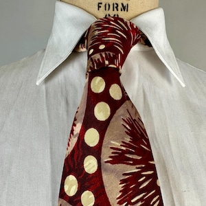 May include: A patterned tie with a white shirt collar. The tie features a design of large cream-coloured circles and abstract shapes in shades of burgundy and tan. The shirt is white.