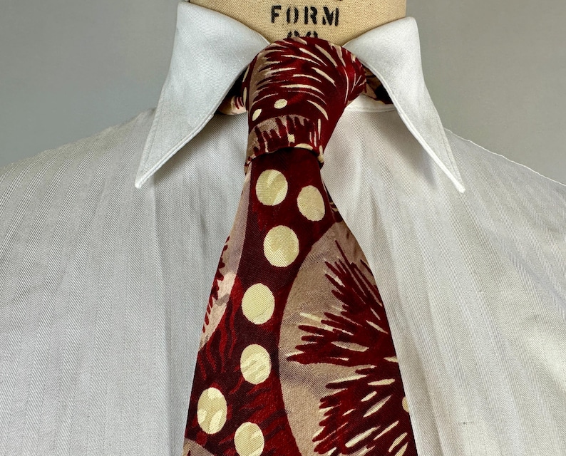 May include: A patterned tie with a white shirt collar. The tie features a design of large cream-coloured circles and abstract shapes in shades of burgundy and tan. The shirt is white.