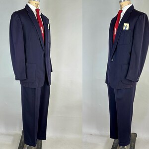 May include: A navy blue suit displayed on a mannequin. The suit includes a blazer with large pockets, a red tie, and matching trousers. A white pocket square is visible in the blazer's breast pocket. The suit is a classic style.