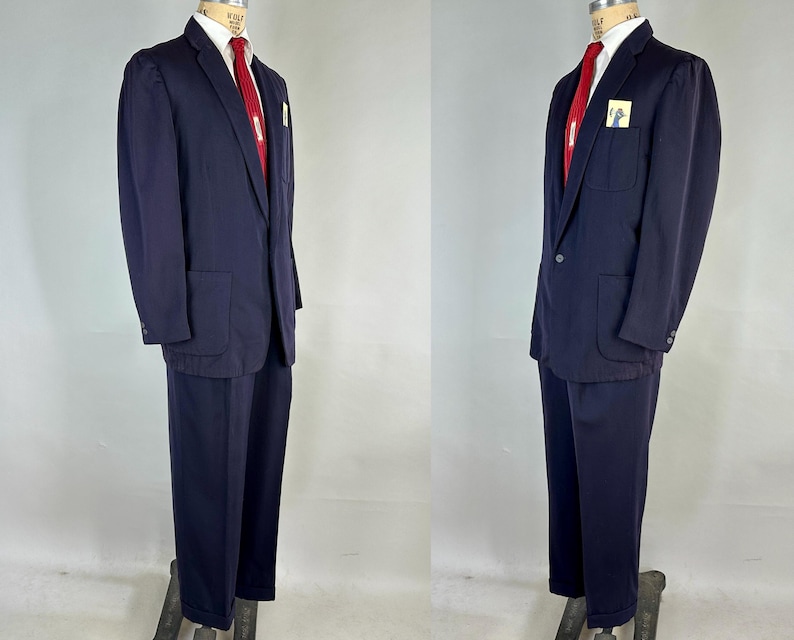 May include: A navy blue suit displayed on a mannequin. The suit includes a blazer with large pockets, a red tie, and matching trousers. A white pocket square is visible in the blazer's breast pocket. The suit is a classic style.