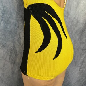 May include: A bright yellow leotard featuring a bold black vertical stripe along the side and a black curved graphic design on the front. The garment is constructed from a ribbed fabric and includes a high-cut leg opening and a scoop neckline.