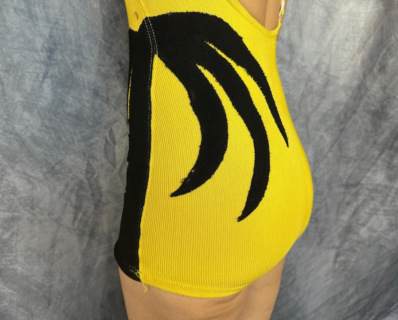 May include: A bright yellow leotard featuring a bold black vertical stripe along the side and a black curved graphic design on the front. The garment is constructed from a ribbed fabric and includes a high-cut leg opening and a scoop neckline.