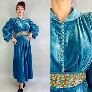 May include: A teal velvet dress with long sleeves and a button-down front. The dress features a beaded belt with colourful accents. The dress has a pleated skirt and is worn with black shoes. The dress is vintage.