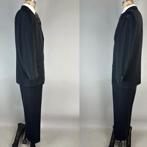 May include: A black suit consisting of a single-breasted blazer with a notched lapel and matching trousers. The blazer features three buttons on each cuff and a white collared shirt underneath with a dark patterned necktie visible at the collar.