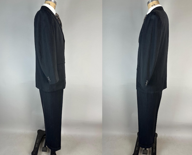 May include: A black suit consisting of a single-breasted blazer with a notched lapel and matching trousers. The blazer features three buttons on each cuff and a white collared shirt underneath with a dark patterned necktie visible at the collar.