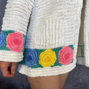 May include: A white textured jacket featuring a horizontal band of raised, tufted floral patterns in shades of pink, yellow, and blue set against a green background. The garment has a ribbed, plush surface texture and long sleeves with matching floral trim.