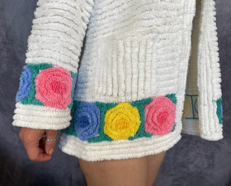 May include: A white textured jacket featuring a horizontal band of raised, tufted floral patterns in shades of pink, yellow, and blue set against a green background. The garment has a ribbed, plush surface texture and long sleeves with matching floral trim.