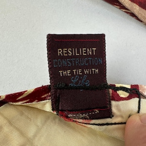 May include: Close-up of a burgundy tie with a cream-coloured lining. The tie's label reads "RESILIENT CONSTRUCTION THE TIE WITH Life" in white and light blue text. The tie has a red and gold floral pattern.