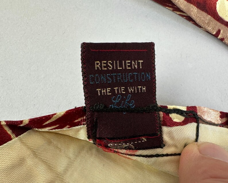 May include: Close-up of a burgundy tie with a cream-coloured lining. The tie's label reads "RESILIENT CONSTRUCTION THE TIE WITH Life" in white and light blue text. The tie has a red and gold floral pattern.