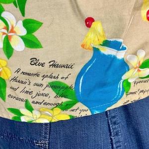 May include: Tan fabric with a tropical print featuring a blue cocktail, yellow pineapple, and red cherries. The design includes white and yellow flowers with green leaves, and the text "Blue Hawaii".