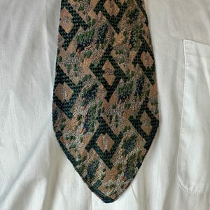May include: A necktie featuring a textured geometric pattern in shades of tan, dark green, and black. The design consists of repeating diamond-like shapes with a mottled, woven appearance, set against a plain white button-down shirt background.
