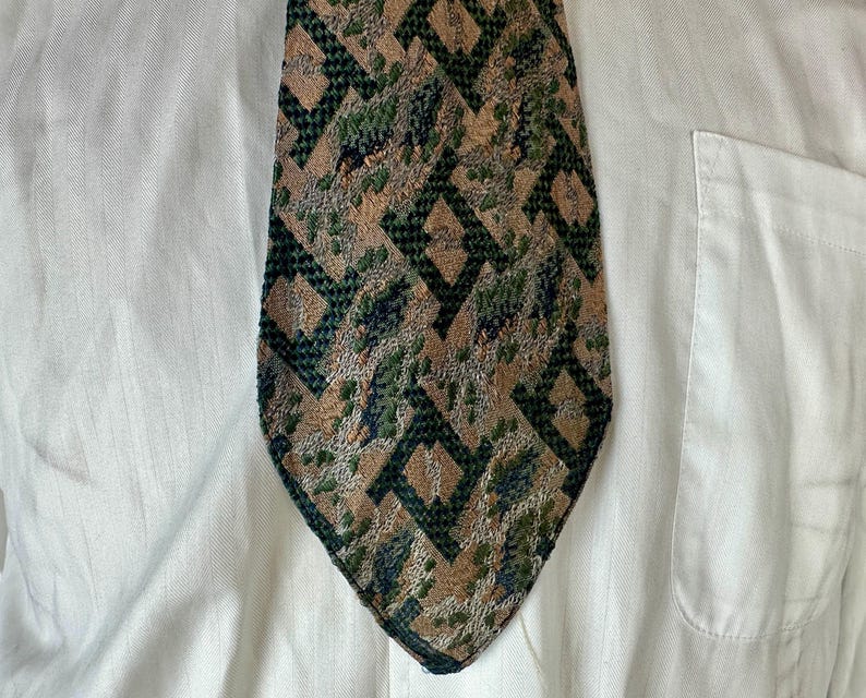 May include: A necktie featuring a textured geometric pattern in shades of tan, dark green, and black. The design consists of repeating diamond-like shapes with a mottled, woven appearance, set against a plain white button-down shirt background.