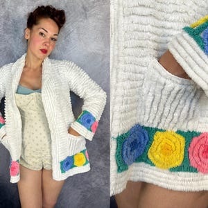 May include: A white textured cardigan featuring long sleeves with blue, yellow, and pink floral-shaped appliques on the cuffs and hem. The garment has a shawl collar, two front pockets, and a soft, ribbed fabric construction throughout.
