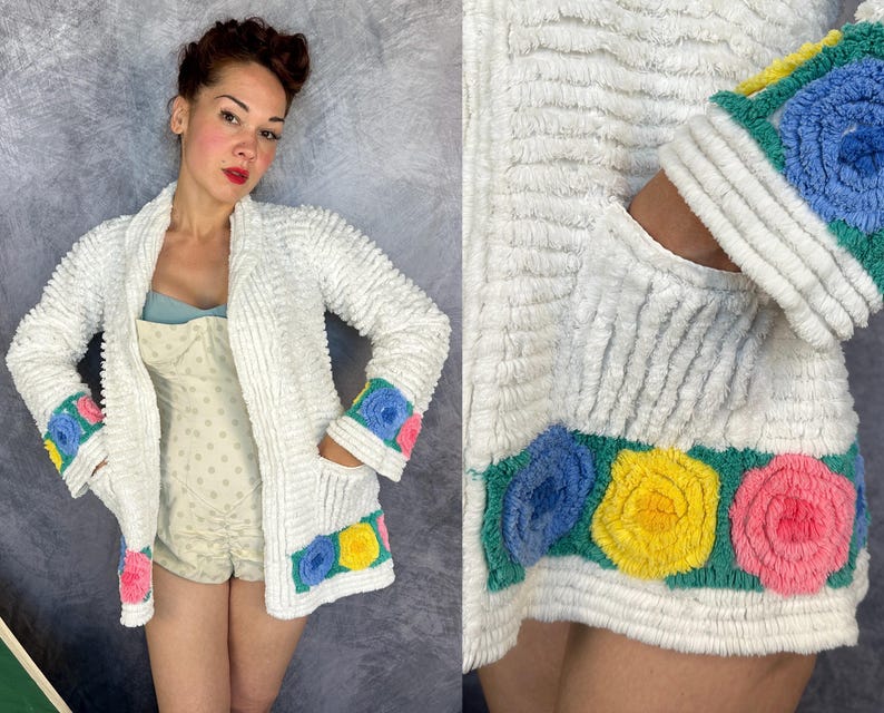 May include: A white textured cardigan featuring long sleeves with blue, yellow, and pink floral-shaped appliques on the cuffs and hem. The garment has a shawl collar, two front pockets, and a soft, ribbed fabric construction throughout.