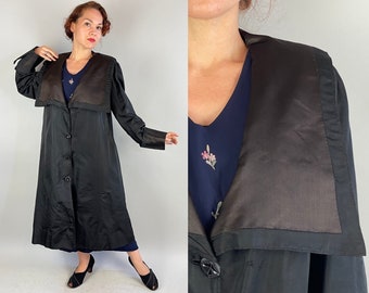 Op de afbeelding: Een lange, zwarte vintage jas met een brede, donkerbruine kraag en een knoopsluiting. De jas heeft lange mouwen en een getailleerde pasvorm. Het model draagt er een donkerblauwe jurk onder. De jas is een klassieke stijl.