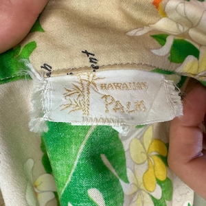 May include: Close-up of a vintage Hawaiian shirt label. The label reads "Hawaiian Palm" with a bamboo illustration. The shirt fabric features a floral pattern with green leaves, white and yellow flowers, and a beige background.