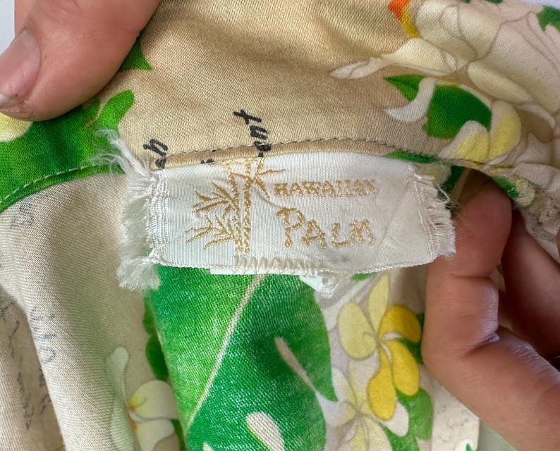 May include: Close-up of a vintage Hawaiian shirt label. The label reads "Hawaiian Palm" with a bamboo illustration. The shirt fabric features a floral pattern with green leaves, white and yellow flowers, and a beige background.