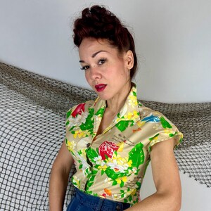 May include: A short-sleeved, button-down shirt with a beige background and a colorful floral and tropical design. The shirt features a collar and is made of a lightweight fabric. The design includes red, yellow, green, and blue elements.