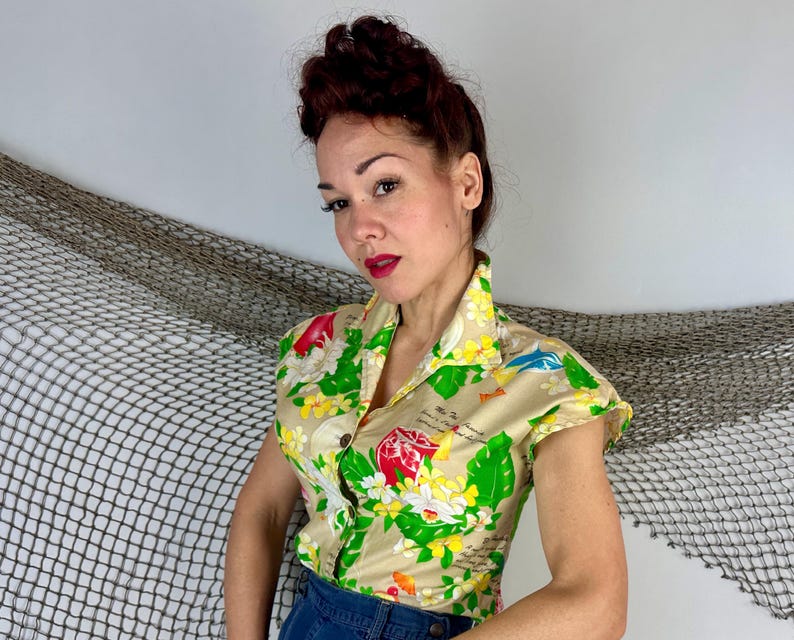 May include: A short-sleeved, button-down shirt with a beige background and a colorful floral and tropical design. The shirt features a collar and is made of a lightweight fabric. The design includes red, yellow, green, and blue elements.