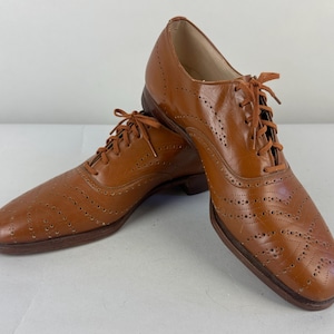 May include: A pair of brown leather Oxford shoes with decorative perforations and matching laces. The shoes have a classic design with a pointed toe and a low heel. The leather has a polished finish.
