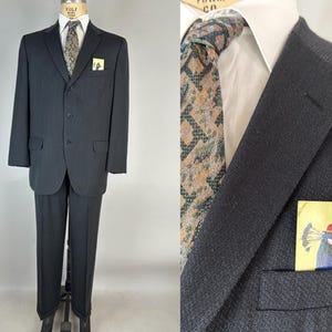 May include: A dark charcoal suit featuring a white collared shirt, a patterned tie in shades of brown and green, and a pocket square displaying a graphic illustration of a peacock wearing a red hat. The suit jacket has three buttons and flap pockets.