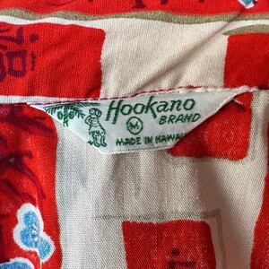 Pode incluir: Uma camisa com padr&atilde;o vermelho e creme apresentando uma etiqueta de tecido branco com texto verde que diz Hookano Brand, M, e Made in Hawaii, acompanhada por um pequeno gr&aacute;fico verde de uma palmeira e uma figura no lado esquerdo.