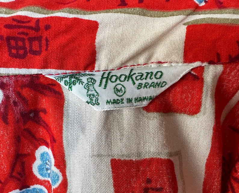 Pode incluir: Uma camisa com padr&atilde;o vermelho e creme apresentando uma etiqueta de tecido branco com texto verde que diz Hookano Brand, M, e Made in Hawaii, acompanhada por um pequeno gr&aacute;fico verde de uma palmeira e uma figura no lado esquerdo.