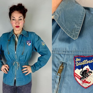 May include: A blue zip-up jacket featuring a drawstring waist, a pointed collar, and multiple embroidered patches. One patch displays the text "les Diablerets" above a graphic of a skier on a mountain. The jacket has two vertical zippered pockets.