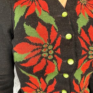 May include: Black cardigan sweater with a poinsettia floral pattern in red and green. The sweater has a V-neckline and is fastened with green buttons. The sleeves are long and the knitwear is visible.