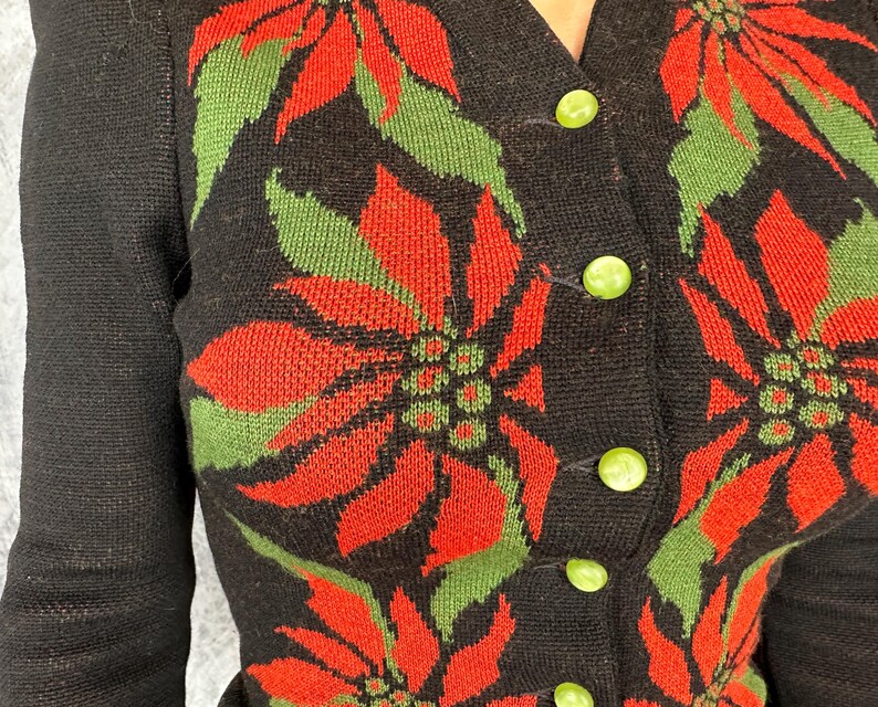 May include: Black cardigan sweater with a poinsettia floral pattern in red and green. The sweater has a V-neckline and is fastened with green buttons. The sleeves are long and the knitwear is visible.