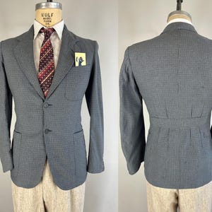 Puede incluir: Una chaqueta de traje gris y negra a cuadros con un cierre de un solo botón. La chaqueta tiene una silueta ajustada y presenta un bolsillo a cada lado. La chaqueta se muestra en un maniquí que lleva una camisa blanca y una corbata roja estampada.