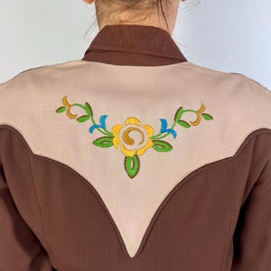 May include: A brown long-sleeve shirt featuring a large, light beige V-shaped yoke on the back. The yoke is decorated with an embroidered floral design in yellow, green, and blue thread, centred across the upper back panel of the garment.