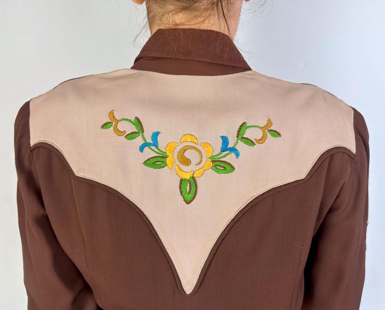 May include: A brown long-sleeve shirt featuring a large, light beige V-shaped yoke on the back. The yoke is decorated with an embroidered floral design in yellow, green, and blue thread, centred across the upper back panel of the garment.