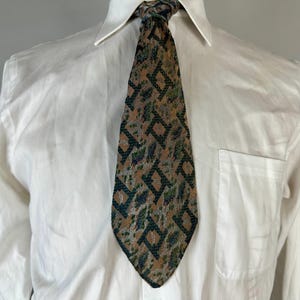 May include: A necktie featuring a tan background with a repeating dark green geometric diamond pattern and mottled grey accents. The tie is worn over a crisp white button-down shirt with a pointed collar and a single chest pocket on the left side.