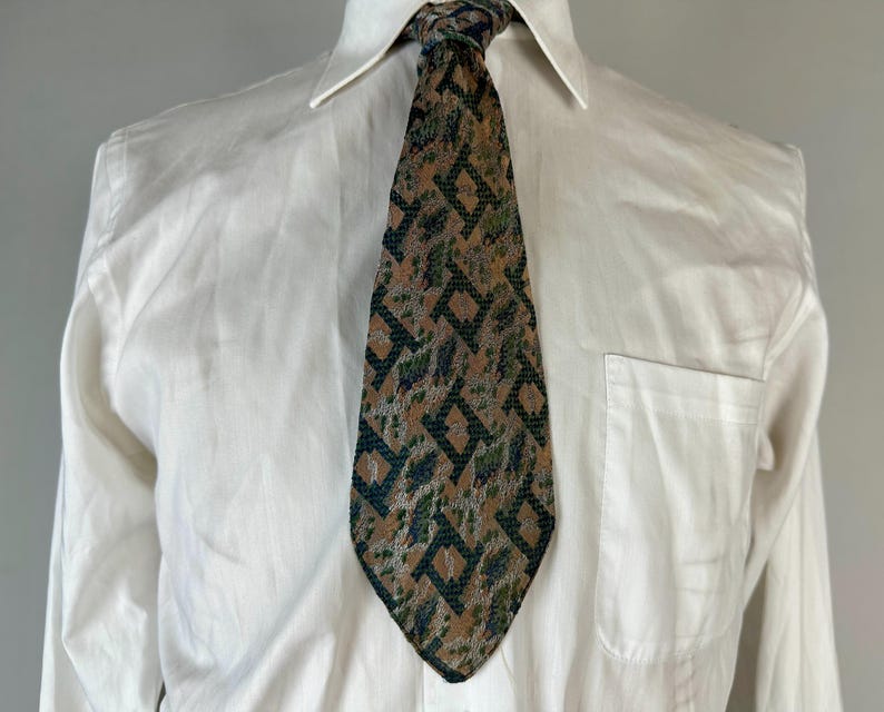 May include: A necktie featuring a tan background with a repeating dark green geometric diamond pattern and mottled grey accents. The tie is worn over a crisp white button-down shirt with a pointed collar and a single chest pocket on the left side.