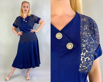 May include: A navy blue dress with a lace overlay on the sleeves and a lace trim at the hem. The dress has a V-neckline and two gold buttons on the front.