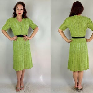 May include: A lime green knitted dress with short sleeves and a V-neck. The dress has a scalloped hemline and is cinched at the waist with a black belt. The dress is knee-length and appears to be vintage.