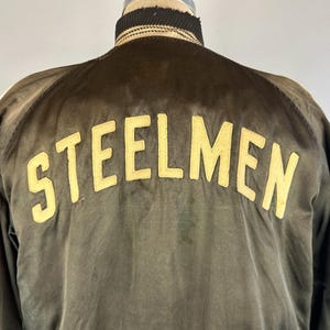 Puede incluir: Una chaqueta bomber de satén verde oscuro con un "STEELMEN" bordado en amarillo en la espalda.