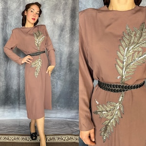 May include: A mauve long-sleeved midi dress featuring a high neckline and shoulder pads. The dress is adorned with a large, vertical gold sequin leaf appliqué extending from the chest to the waist, cinched by a thin black braided belt.