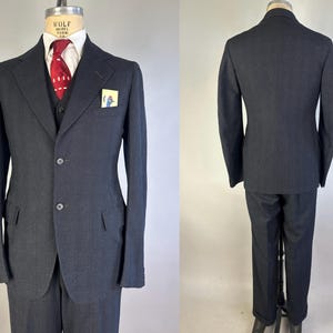 May include: A charcoal gray three-piece suit featuring a two-button jacket with notched lapels, a matching vest, and trousers. The ensemble includes a white collared shirt, a red patterned necktie, and a small pocket square with a bird graphic.