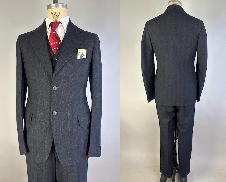 May include: A charcoal gray three-piece suit featuring a two-button jacket with notched lapels, a matching vest, and trousers. The ensemble includes a white collared shirt, a red patterned necktie, and a small pocket square with a bird graphic.