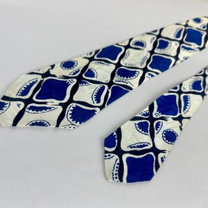 May include: A necktie featuring a repeating geometric pattern of deep blue squares with rounded corners and scalloped edges set against a cream-colored background. The design includes dark blue intersecting lines that create a grid-like structure.